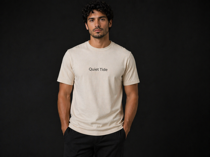 Handsome male model wearing Quiet Tide essential t-shirt in natural color with minimalist logo on front against dark background