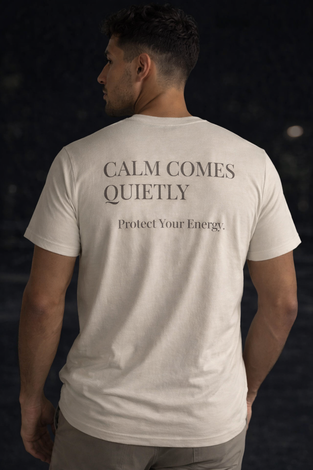 Man wearing Quiet Tide natural t-shirt back view with “Calm Comes Quietly Protect Your Energy” text on dark background